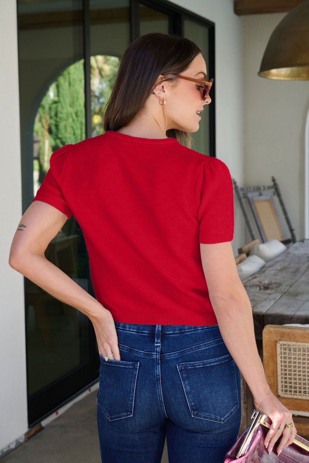 A woman in the Maura Red Short Sleeve Sweater Cardigan stands outdoors with a pink purse and patio furniture nearby.