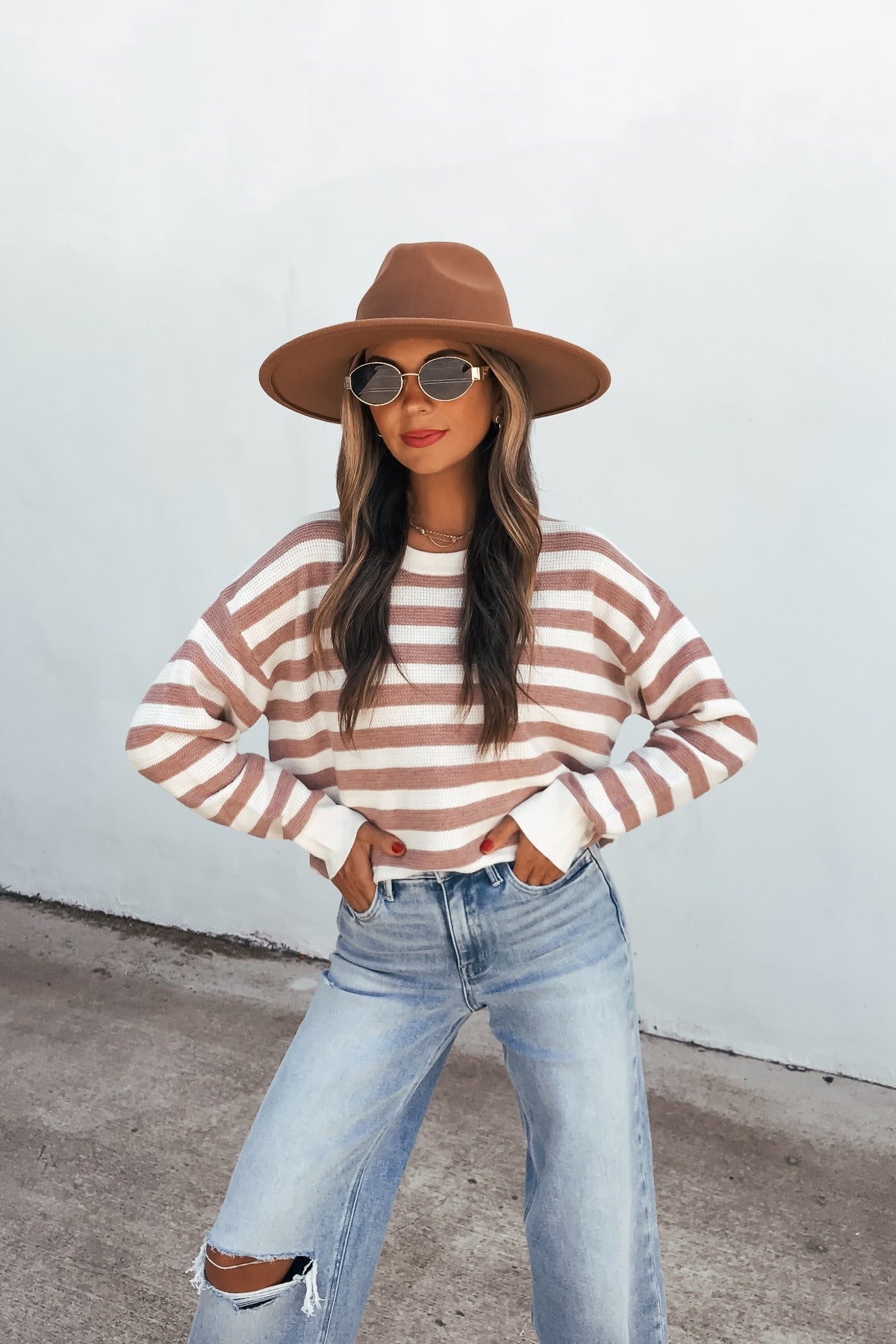 Woman in mauve striped waffle knit sweater, wide-brim hat, sunglasses, and ripped jeans stands against white wall, hands in her pockets.