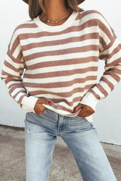 Woman in a mauve striped waffle knit sweater, light blue jeans, and a necklace, standing with hands in pockets.