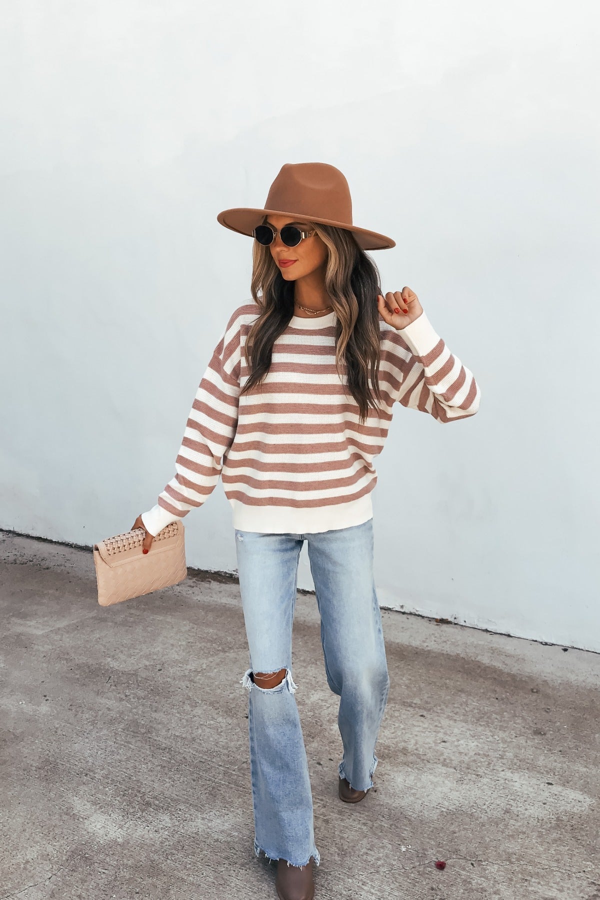 Woman in a brown hat, Mauve Striped Waffle Knit Sweater, jeans, and boots holds a beige handbag while walking by a white wall.