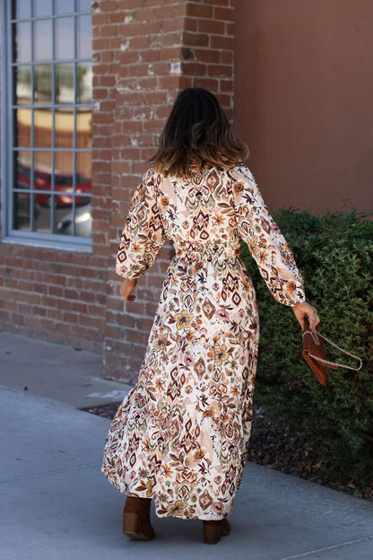A woman in the Meadow Brown Print Tiered Maxi Dress - FINAL SALE and brown boots walks on a sidewalk by a brick wall and green shrub.