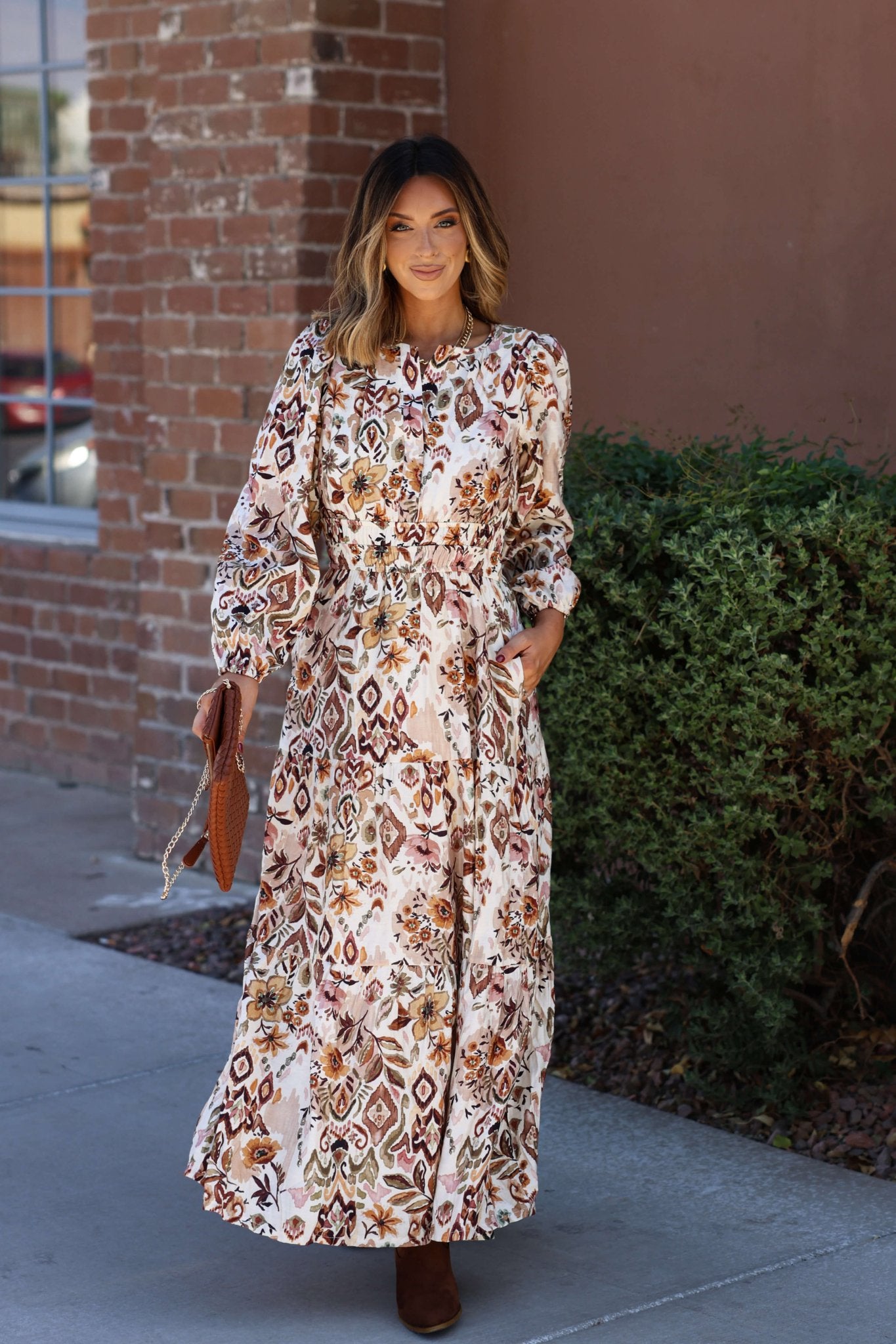 A woman stands outside by a brick wall in the Meadow Brown Print Tiered Maxi Dress - FINAL SALE, styled with brown boots and a purse.