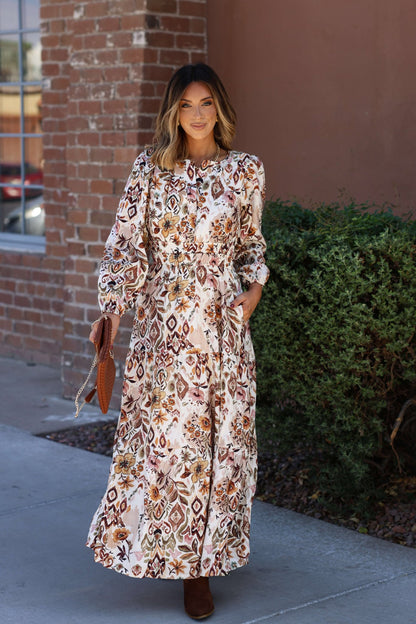 A woman stands outside by a brick wall in the Meadow Brown Print Tiered Maxi Dress - FINAL SALE, styled with brown boots and a purse.