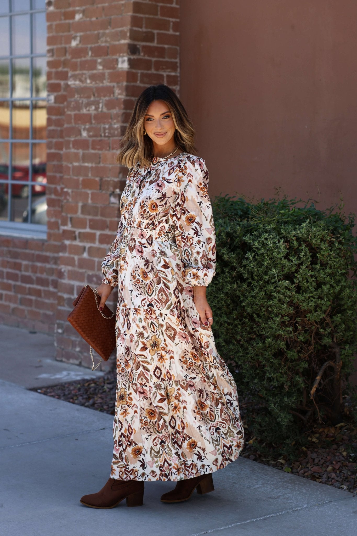 Woman wearing the Meadow Brown Print Tiered Maxi Dress - FINAL SALE stands by a brick wall, smiling and holding a clutch.
