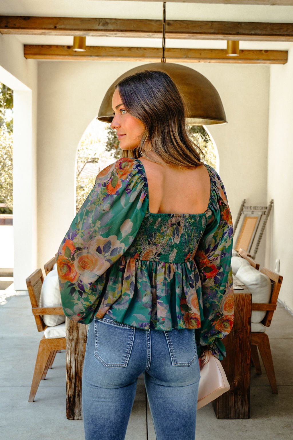 A woman in blue jeans and a Meadow Green Floral Print Smocked Top stands indoors, facing away with light brown hair.