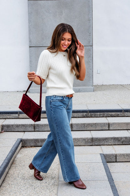 Woman in a white sweater and Medium Wash Baggy Wide Leg Jeans walks outside, carrying a burgundy purse and smiling.