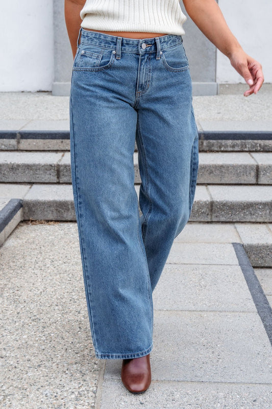 Person wears Medium Wash Baggy Wide Leg Jeans, a white top, and brown shoes while walking on outdoor steps.