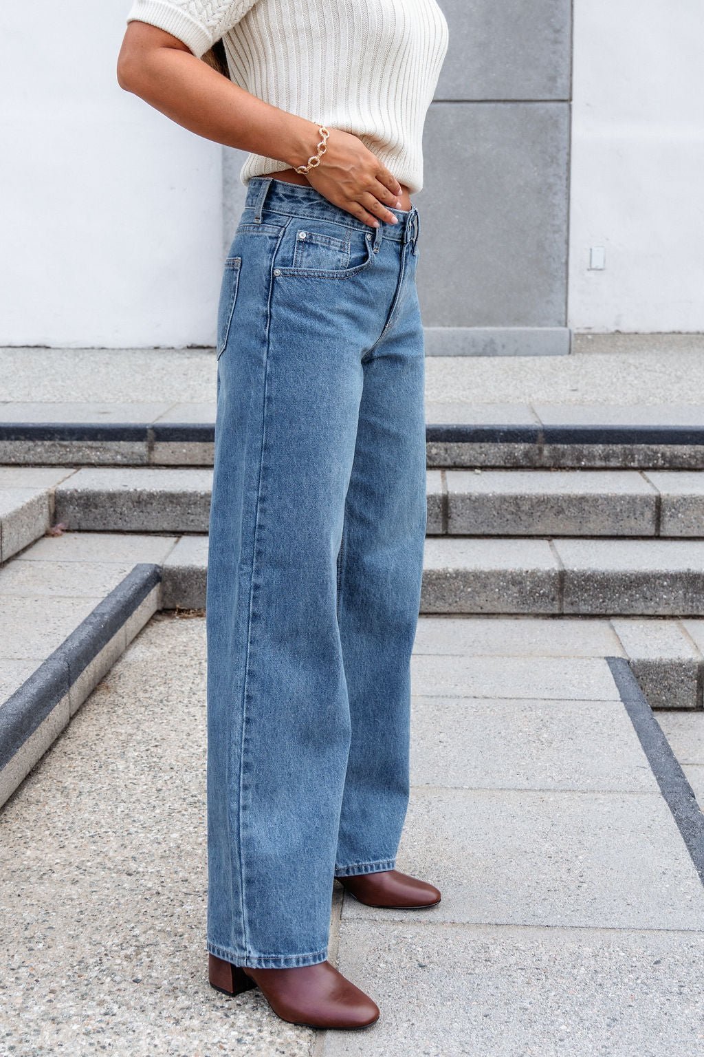 Person wearing a cream knit top, Medium Wash Baggy Wide Leg Jeans, and brown boots, standing outdoors on concrete steps.