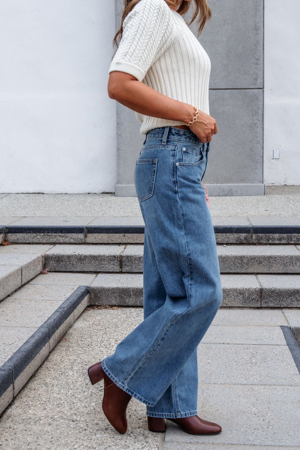A woman in a white knit top and Medium Wash Baggy Wide Leg Jeans with brown heeled boots stands near outdoor steps.