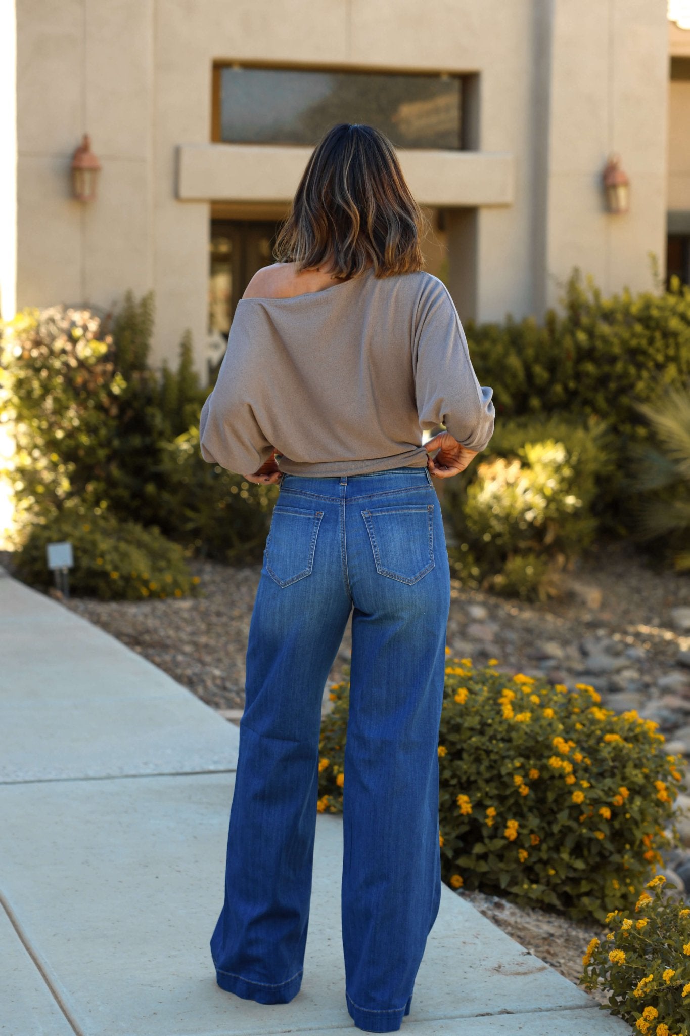 A woman with wavy hair stands outside in a loose taupe top and Medium Wash High Rise Pull-On Wide Leg Jeans.