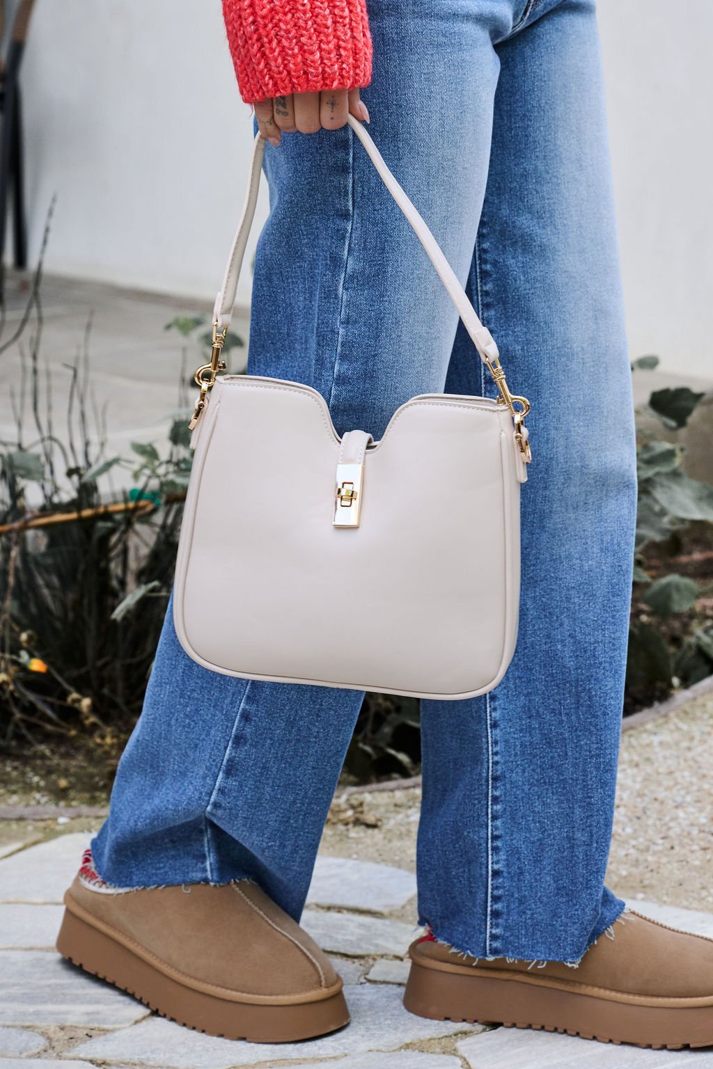 Person in blue jeans and tan shoes holds the Melissa Oatmeal Vegan Leather Shoulder Bag with gold clasp on a stone path.