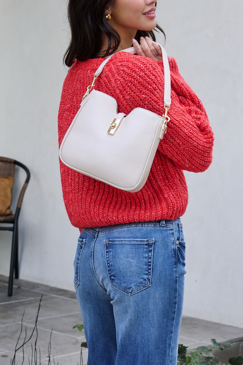 Woman in a red sweater and blue jeans holds the Melissa Oatmeal Vegan Leather Shoulder Bag indoors near a chair.