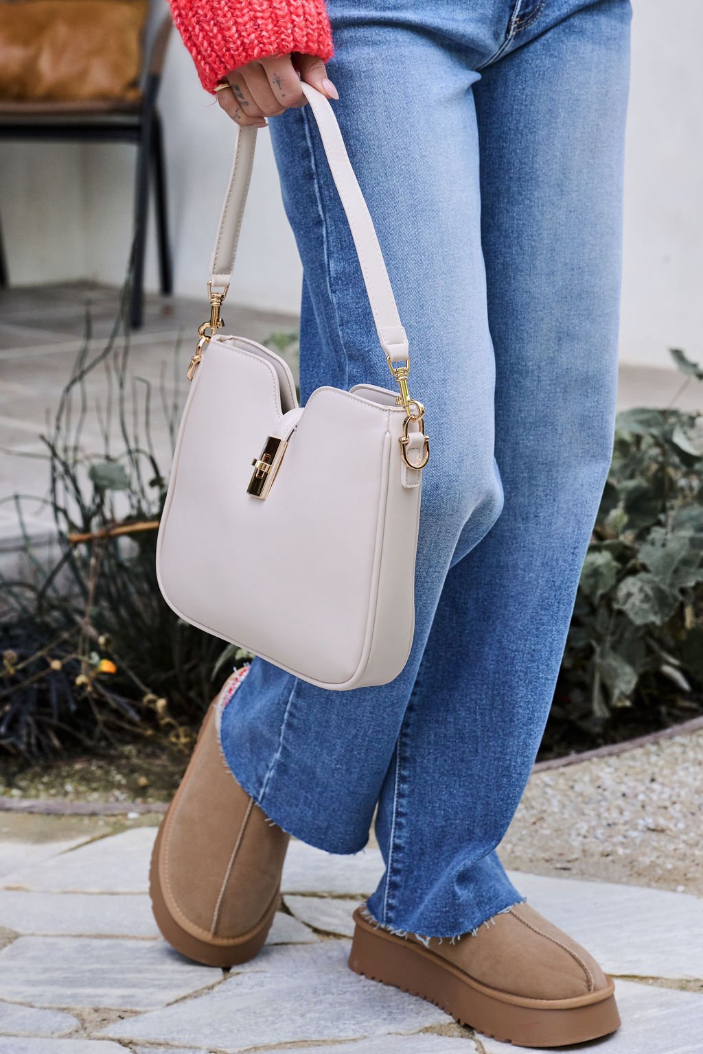 A person in blue jeans and tan shoes holds the Melissa Oatmeal Vegan Leather Shoulder Bag, standing on a stone path.