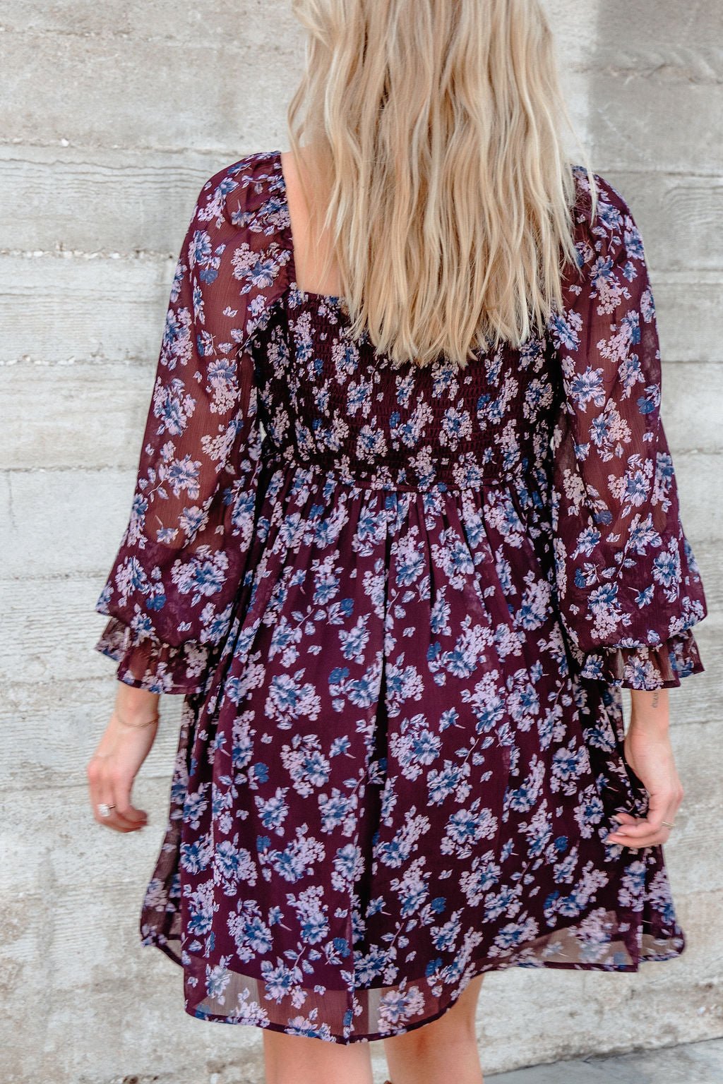 A woman with long blonde hair wears the Merlot Floral Chiffon Babydoll Mini Dress, standing by a textured concrete wall.