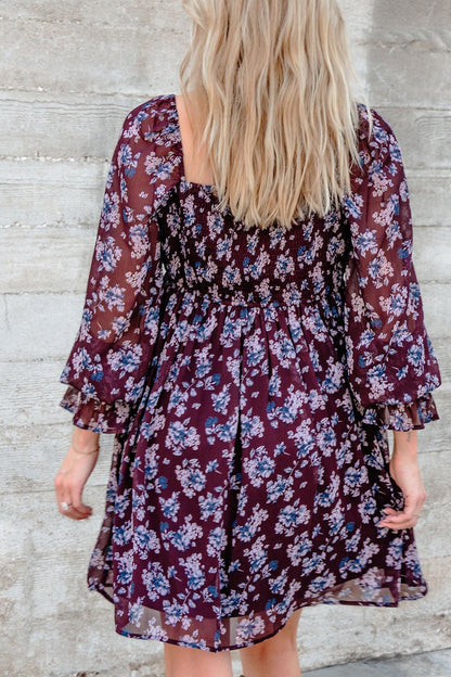 A woman with long blonde hair wears the Merlot Floral Chiffon Babydoll Mini Dress, standing by a textured concrete wall.
