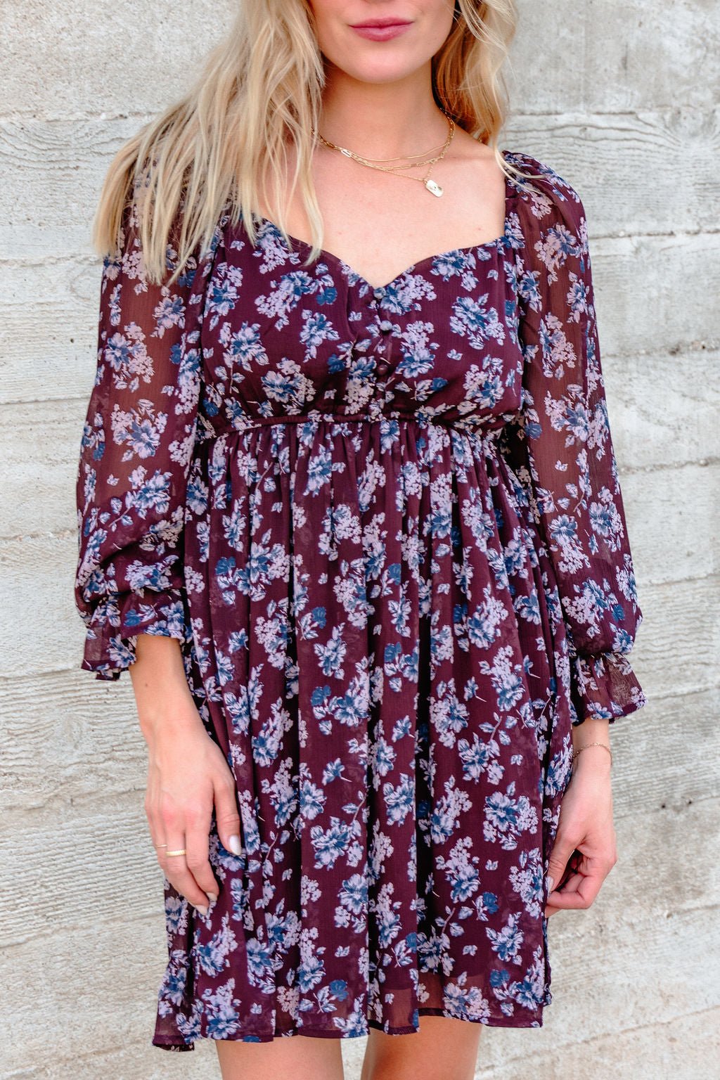 Woman in a Merlot Floral Chiffon Babydoll Mini Dress stands by a textured concrete wall.