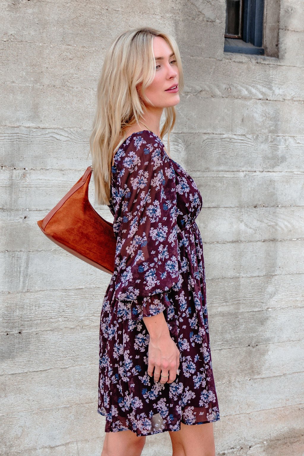A woman in the Merlot Floral Chiffon Babydoll Mini Dress stands sideways by a concrete wall, holding a brown shoulder bag.