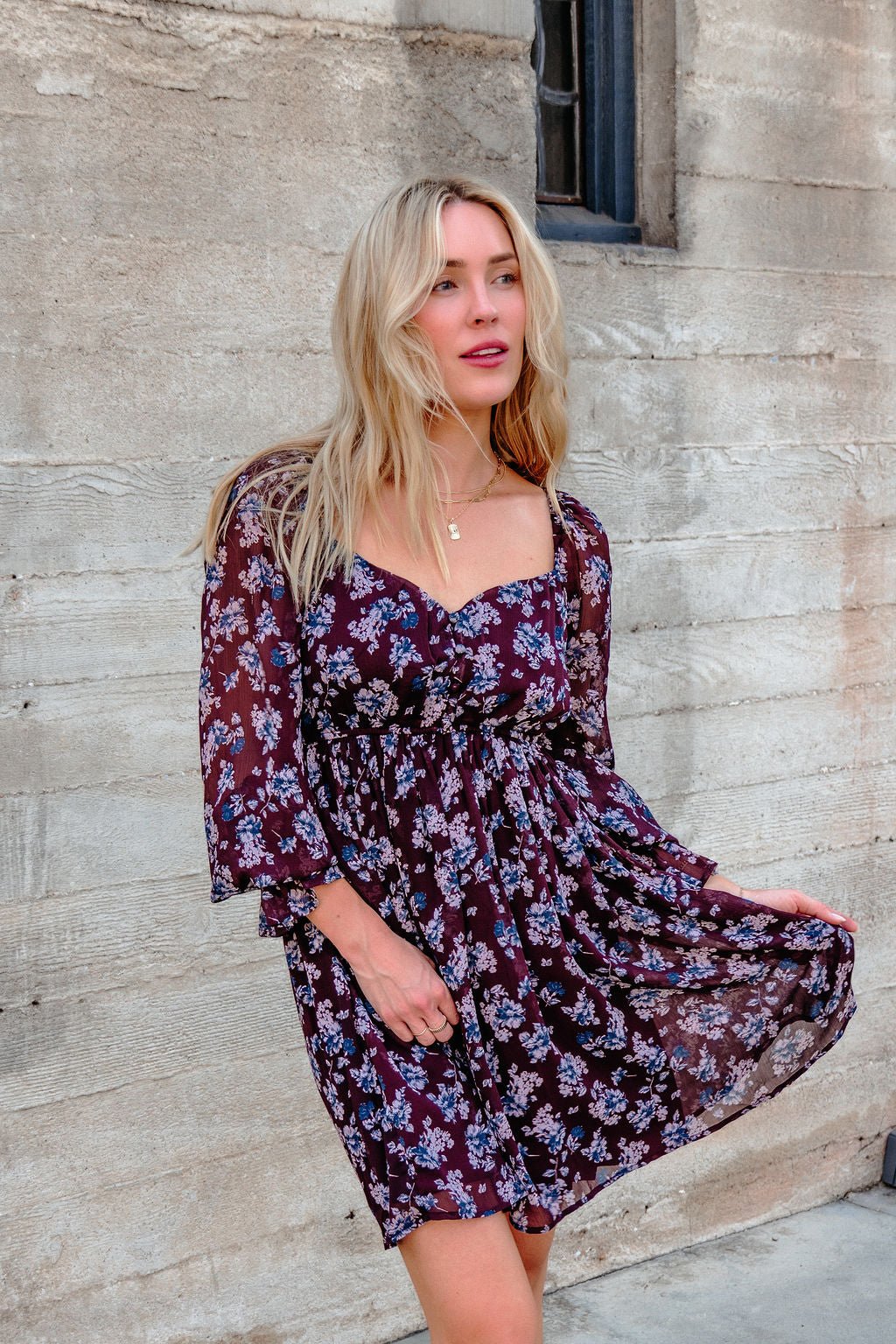 Woman in the Merlot Floral Chiffon Babydoll Mini Dress stands by a textured beige wall, holding her dress and looking to the left.
