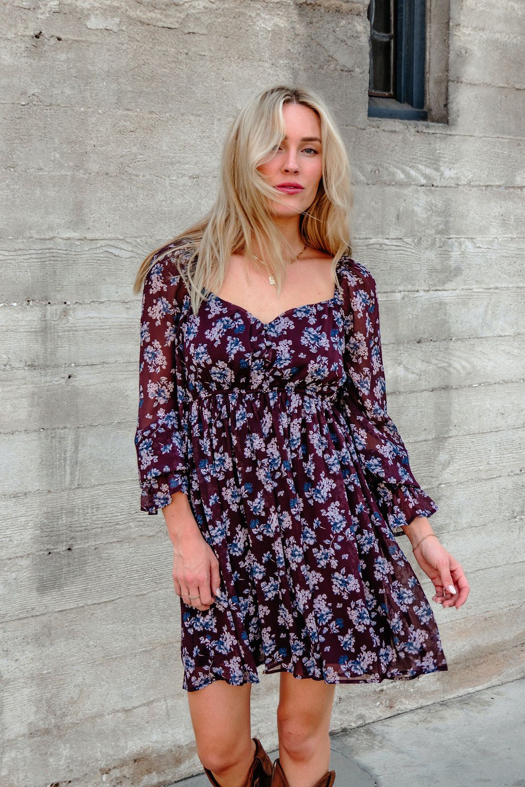 A woman in the Merlot Floral Chiffon Babydoll Mini Dress stands by a textured wall, gazing at the camera with loose blonde hair.
