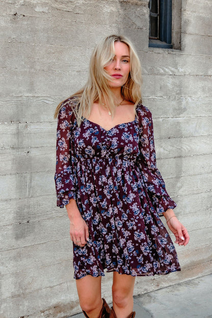 A woman in the Merlot Floral Chiffon Babydoll Mini Dress stands by a textured wall, gazing at the camera with loose blonde hair.