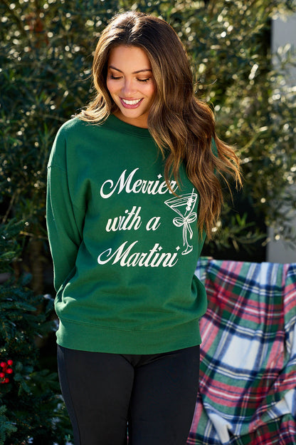 A woman relaxes outdoors by a plaid blanket in the green Merrier With A Martini Graphic Sweatshirt.