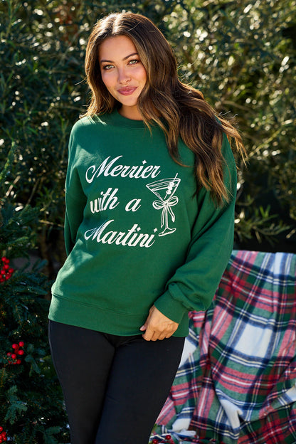 Woman wearing the green Merrier With A Martini Graphic Sweatshirt, outdoors by plaid blanket and greenery.