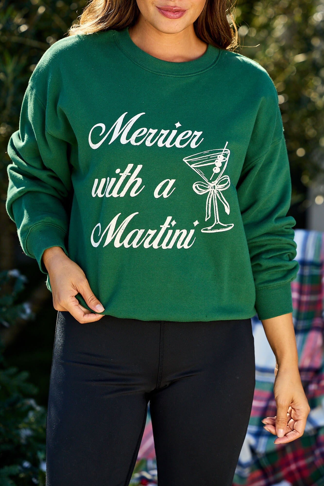 Woman in a green "Merrier With A Martini" graphic sweatshirt standing outdoors.