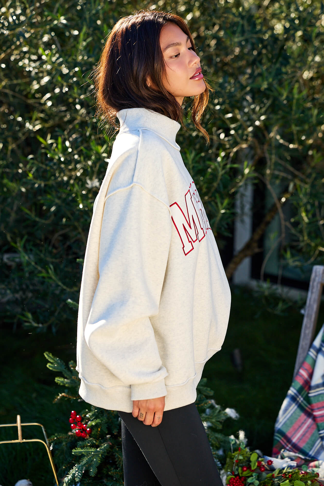 A woman wears the Merry Grey Oversized Sweatshirt - FINAL SALE, standing outdoors near holiday greenery and looking to the side.