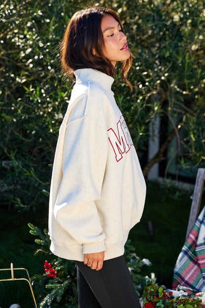 A woman wears the Merry Grey Oversized Sweatshirt - FINAL SALE, standing outdoors near holiday greenery and looking to the side.