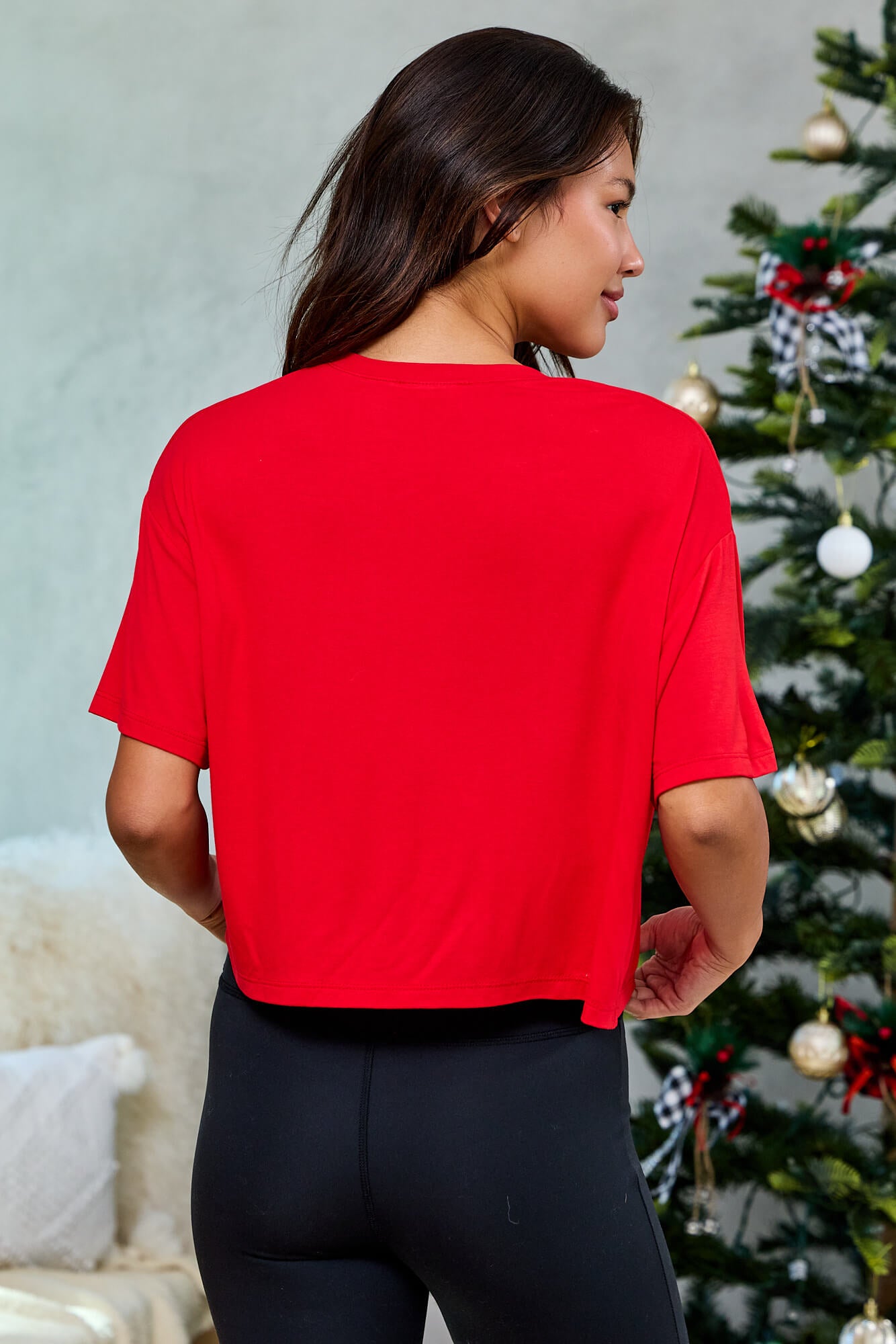 Woman in a red "Merry" Mood Graphic Tee (Final Sale) and black pants stands by a decorated Christmas tree, facing away.