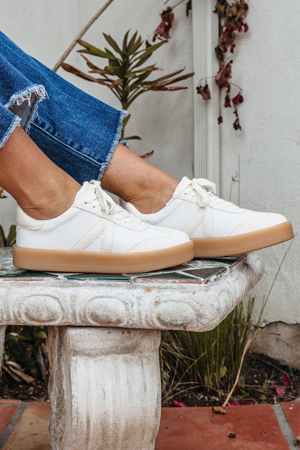 Person in Mia Axel Natural Lace Up Sneakers, gum soles, blue jeans, seated on a stone bench near lush green plants outdoors.