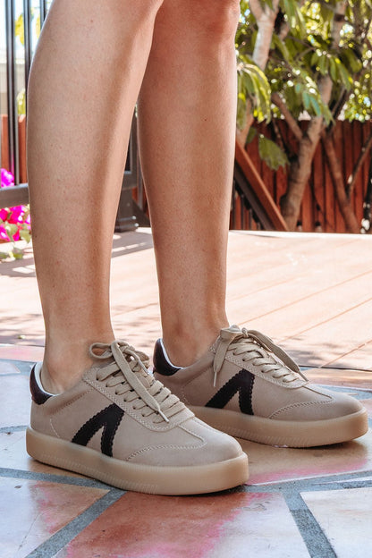 A person wears Mia Axel Taupe Lace Up Sneakers - FINAL SALE on a tiled patio, surrounded by lush plants.