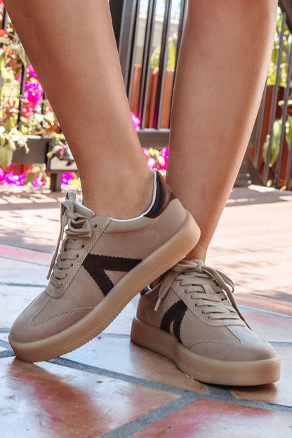 Close-up of a person wearing Mia Axel Taupe Lace Up Sneakers - FINAL SALE, standing on colorful tiles with pink flowers behind.