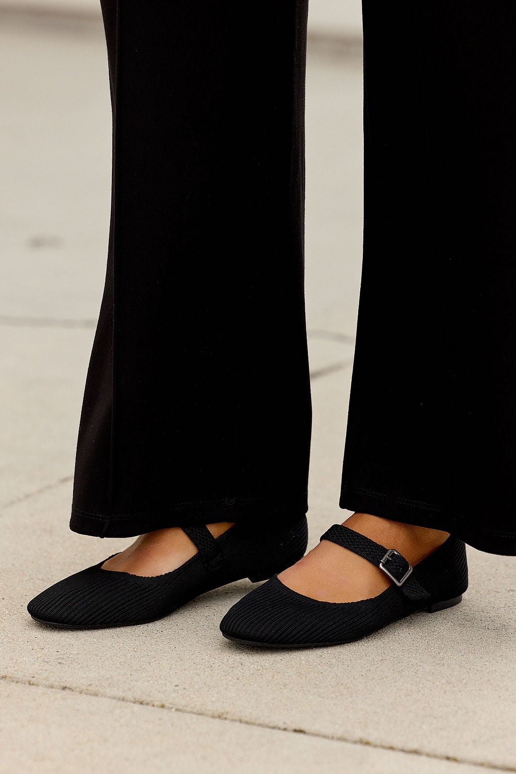 Someone wearing MIA Brita Ballet Flats - Black and black pants stands on light-colored pavement.