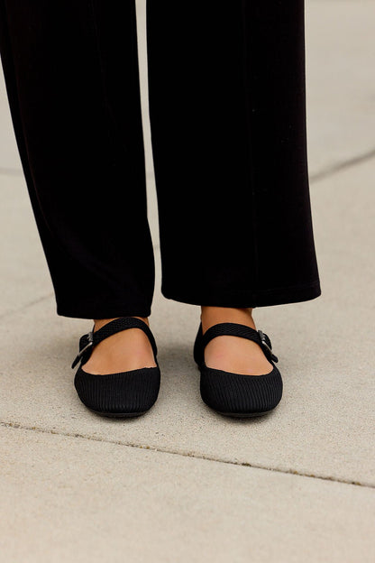 Person wearing black pants and MIA Brita Ballet Flats in Black with a Mary Jane strap, standing on a light concrete surface.