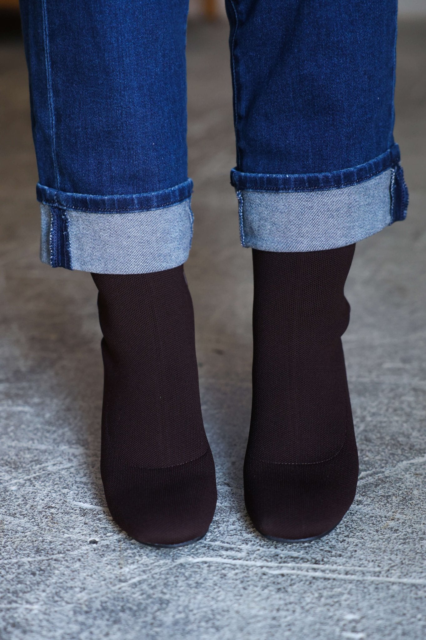 Person wears MIA Bronwen Brown Heeled Ankle Booties with rolled-up blue jeans, standing on a textured gray surface.
