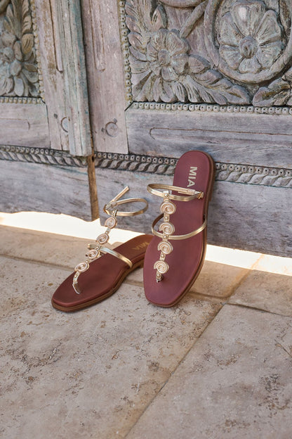 MIA Dorcie Strappy Sandals in gold with adjustable ankle straps and maroon soles, shown on stone before an ornate wooden door.