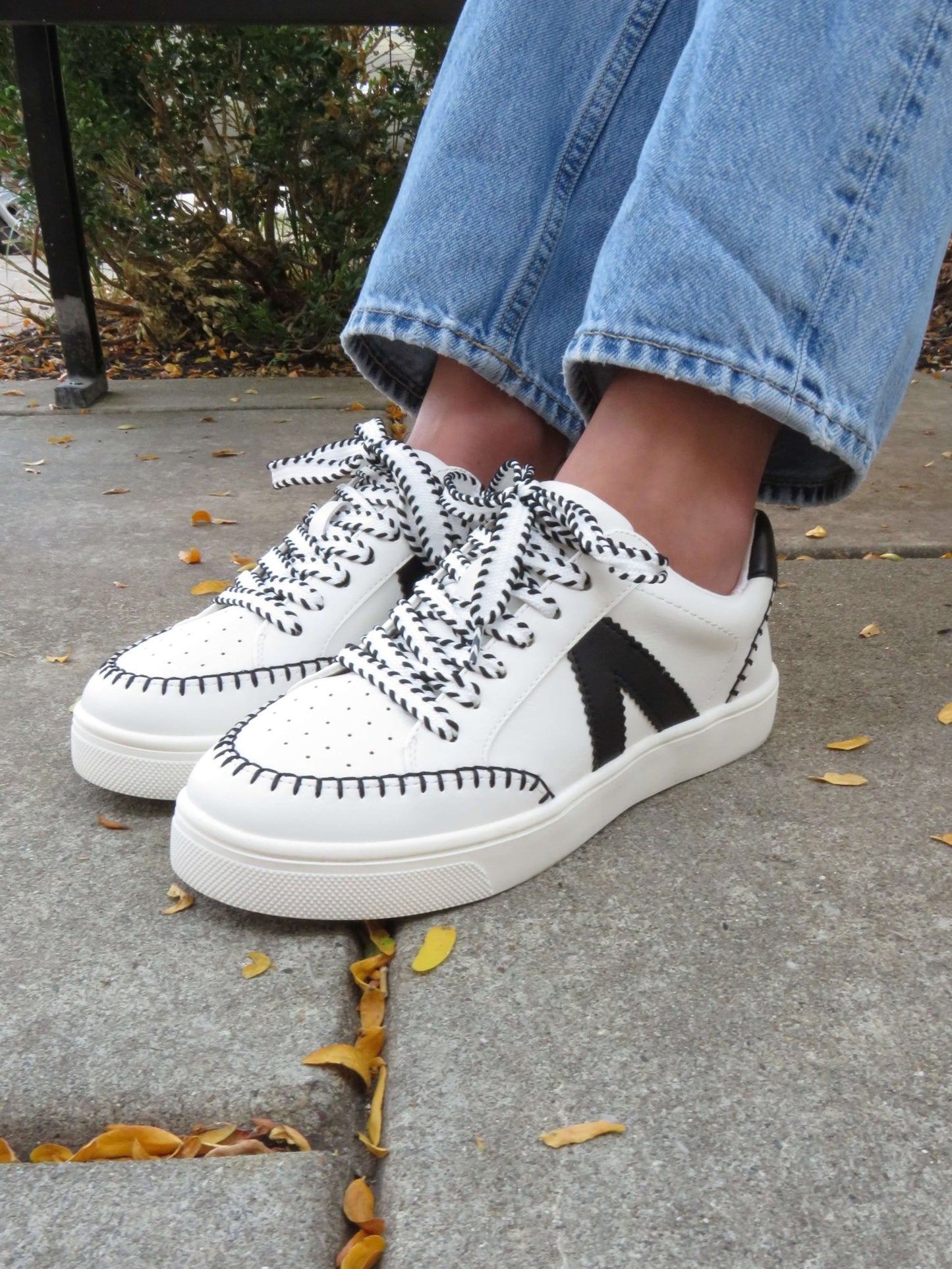 MIA Itzia Black and White Stitched Sneakers pair well with light blue wide-leg jeans for a comfy, casual look among yellow leaves.
