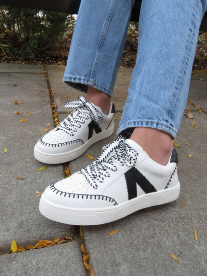 Person wears MIA Itzia Black and White Stitched Sneakers with blue jeans while walking on a leafy sidewalk.