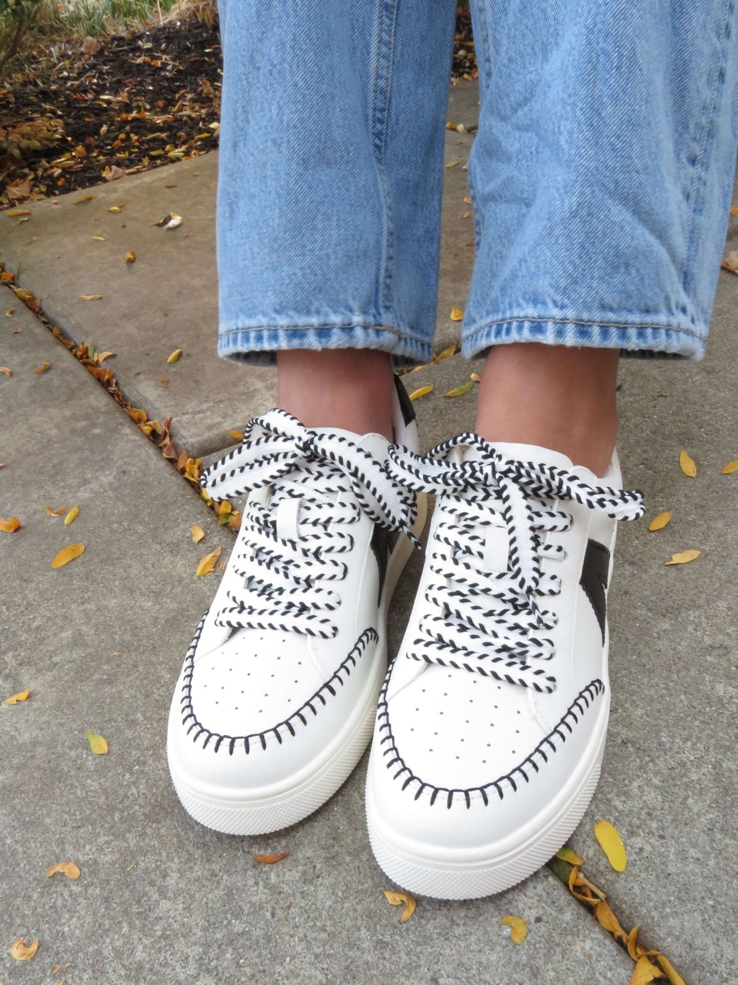 Someone in light blue jeans and MIA Itzia Black and White Stitched Sneakers stands on a sidewalk with fallen leaves.