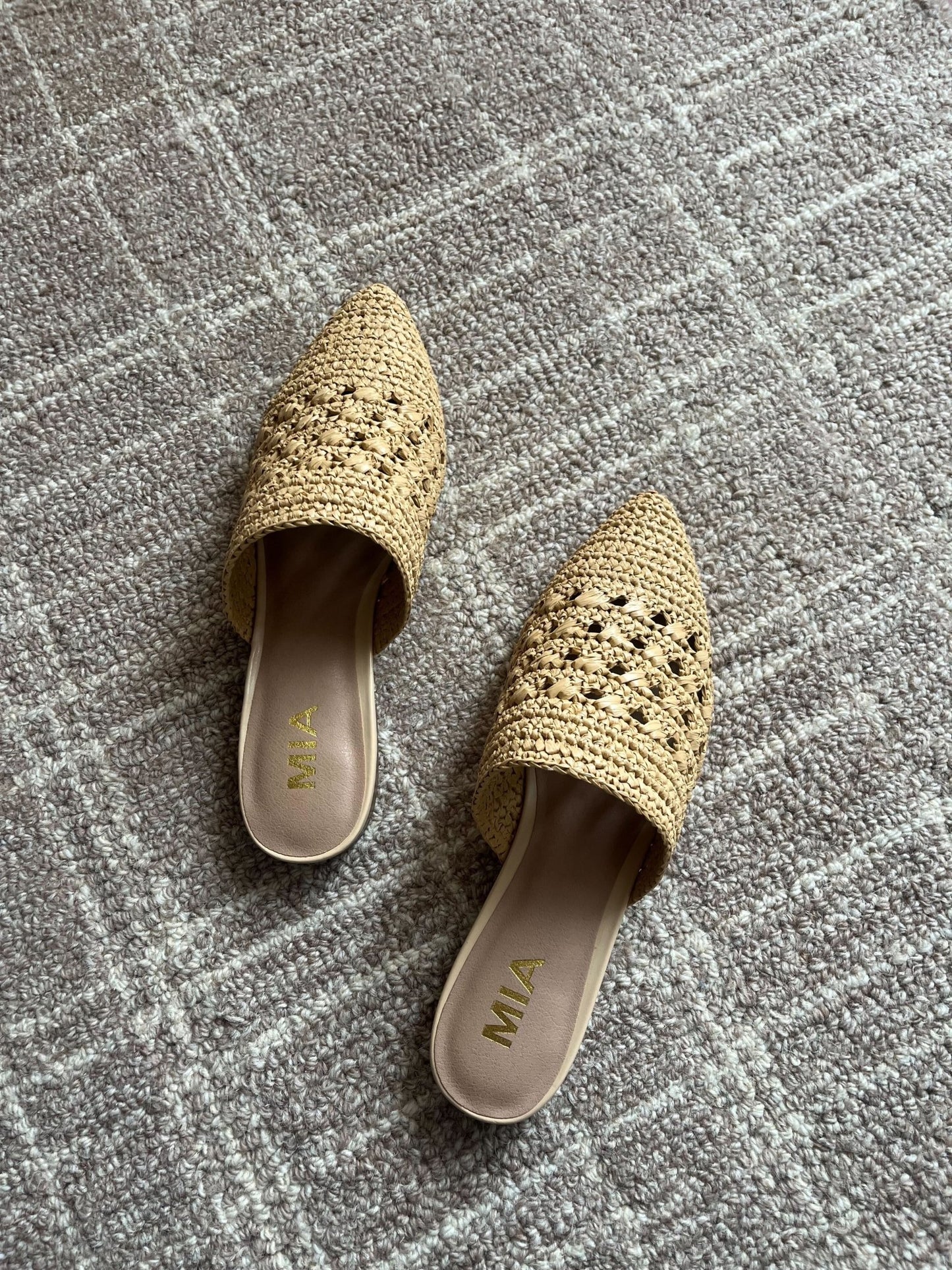 The MIA Jany Natural Pointed Mules are woven, tan-colored shoes with pointed toes, displayed on a textured gray carpet.