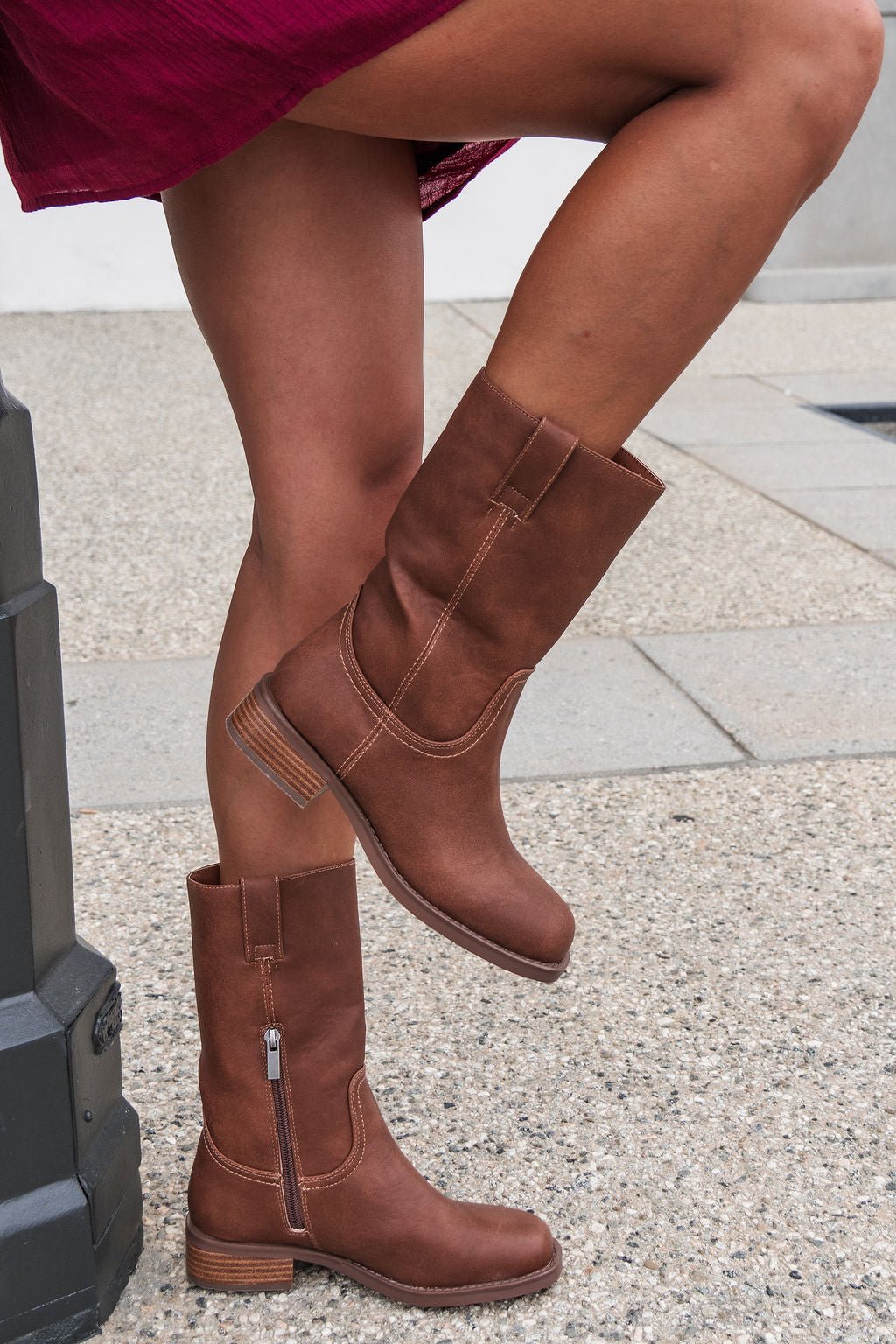 Someone in MIA Miley Mid Calf Boots - Tan balances on one leg outdoors on a stone sidewalk.