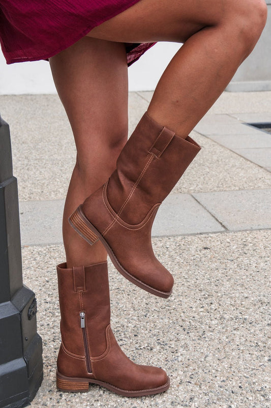 Someone in MIA Miley Mid Calf Boots - Tan balances on one leg outdoors on a stone sidewalk.
