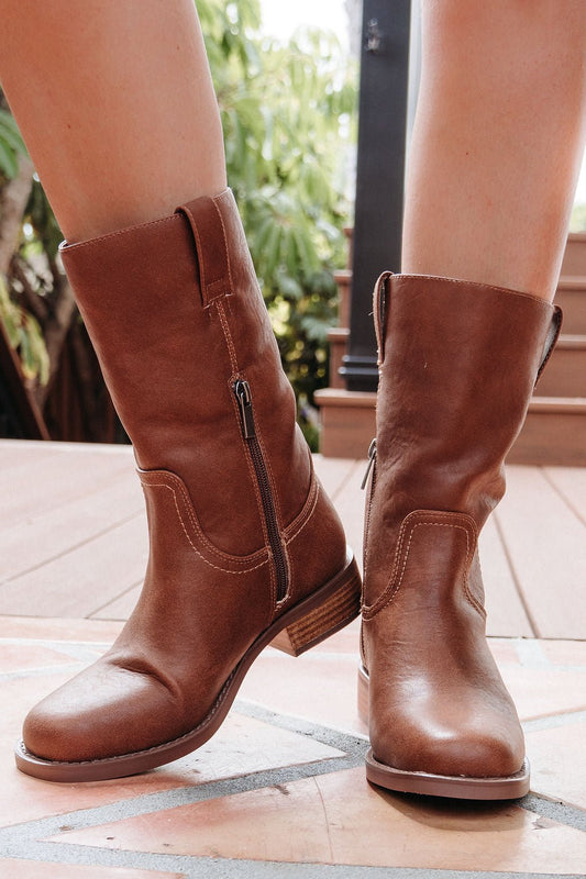 A person wears MIA Miley Mid Calf Boots in tan with side zippers while standing on a wooden deck.