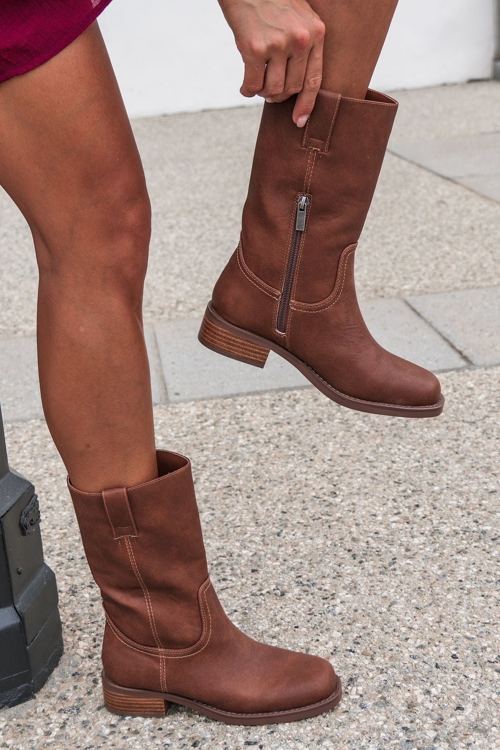 Someone in MIA Miley Mid Calf Boots - Tan stands on a sidewalk, holding up one boot to show its side zipper.