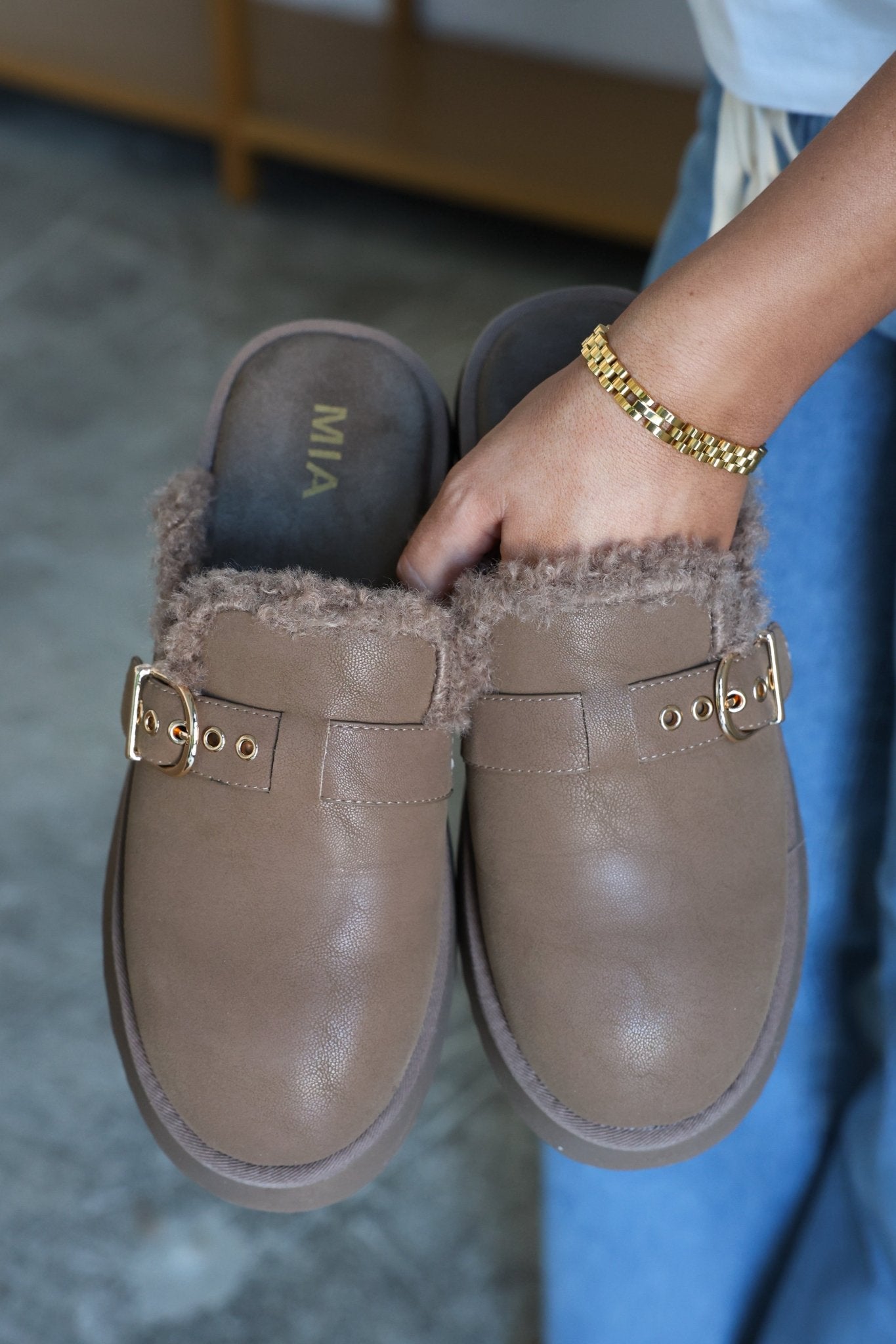 A person holds a pair of MIA Sugary Taupe Sherpa Mules with gold buckles and wears a gold bracelet.