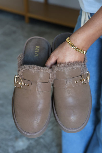 A person holds a pair of MIA Sugary Taupe Sherpa Mules with gold buckles and wears a gold bracelet.