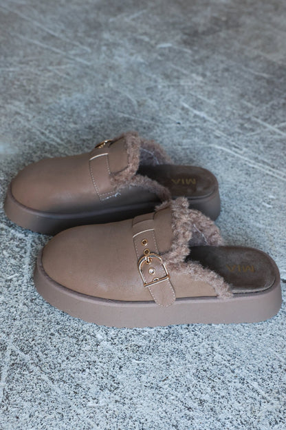 The MIA Sugary Taupe Sherpa Mule features brown faux shearling, gold buckles, and sits on a textured gray surface.