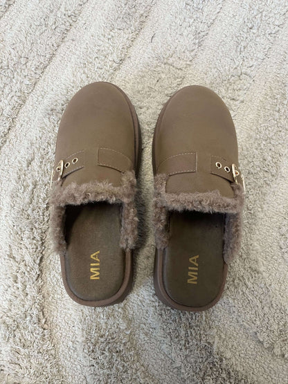 The MIA Sugary Taupe Sherpa Mule features gold buckles and faux fur lining, displayed on a beige textured rug.