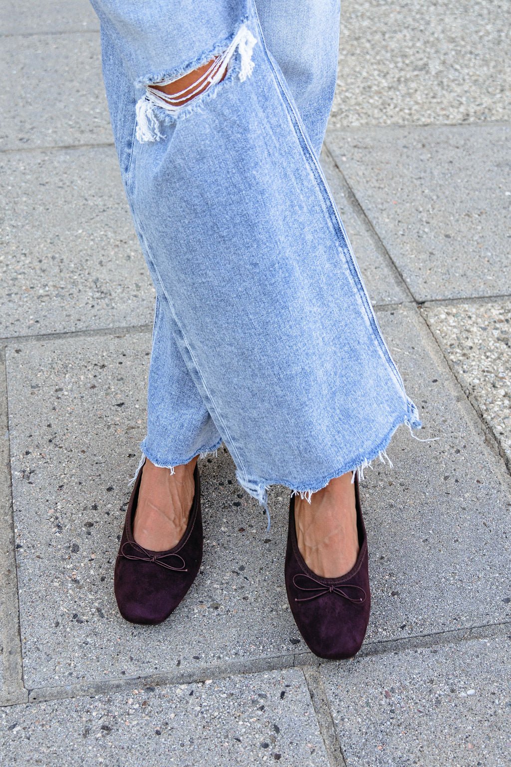 Person wears ripped, light-wash jeans and MIA Tinna Bow Ballet Flats in brown, standing on a sidewalk with crossed feet.